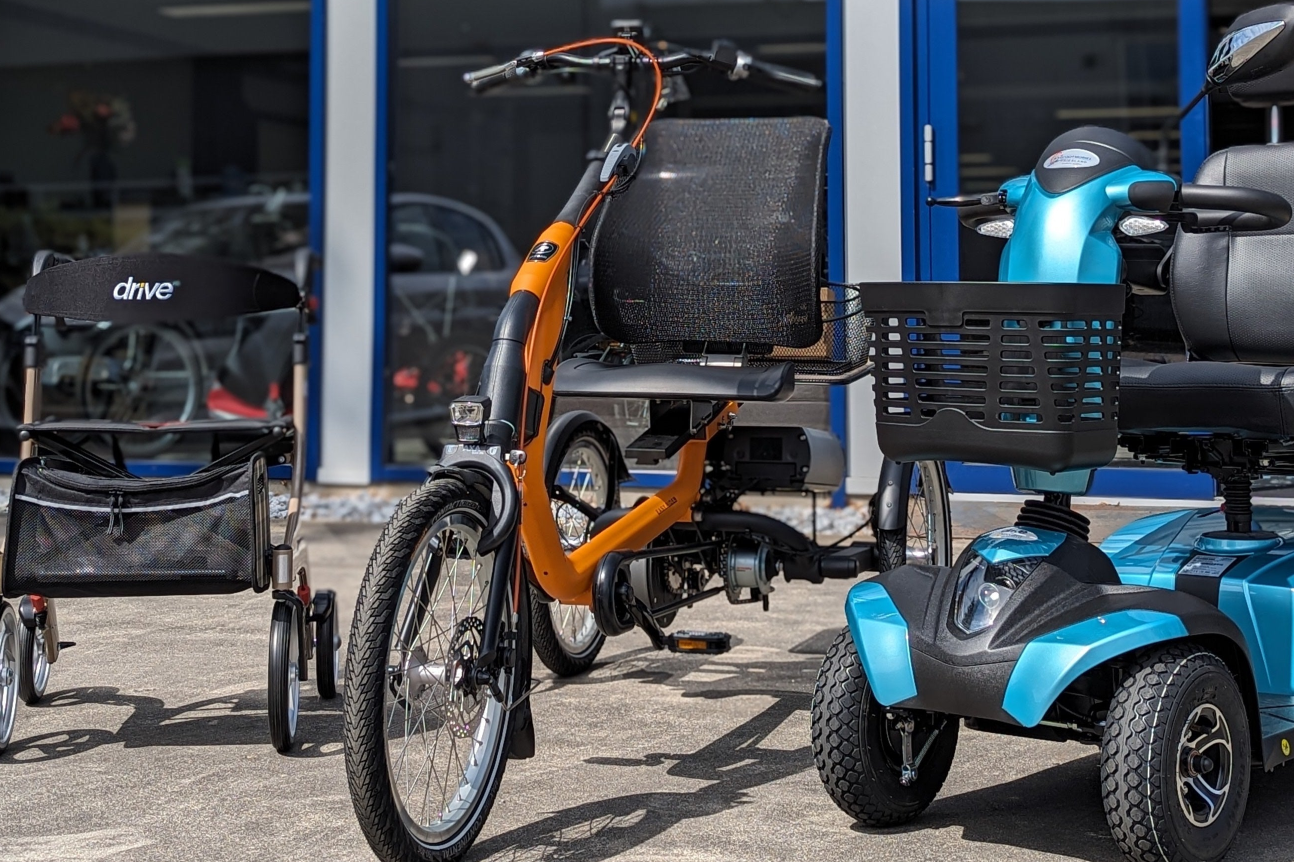 Op deze foto staan de drie hoofdproducten naast elkaar, een oranje aangepaste driewielfiets in het midden, links op de afbeelding de rollator en rechts van de afbeelding een blauwe scootmobiel. Ze staan voor de blauwe showroom.